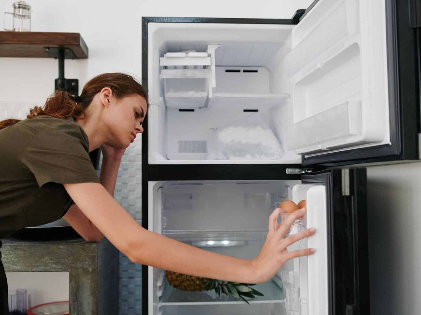 Frustrated woman inspecting a refrigerator freezer with frost buildup, checking why the fridge isn’t cooling.
